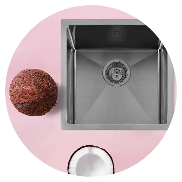 Stainless steel kitchen sink with a whole coconut and half coconut on pink circular background.