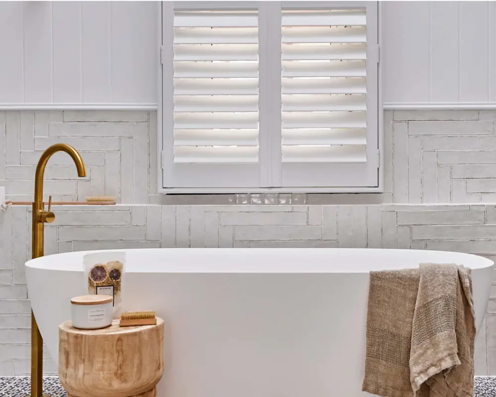 Modern white bathtub with brass faucet, wooden stool with bath products, and white plantation shutters on window.