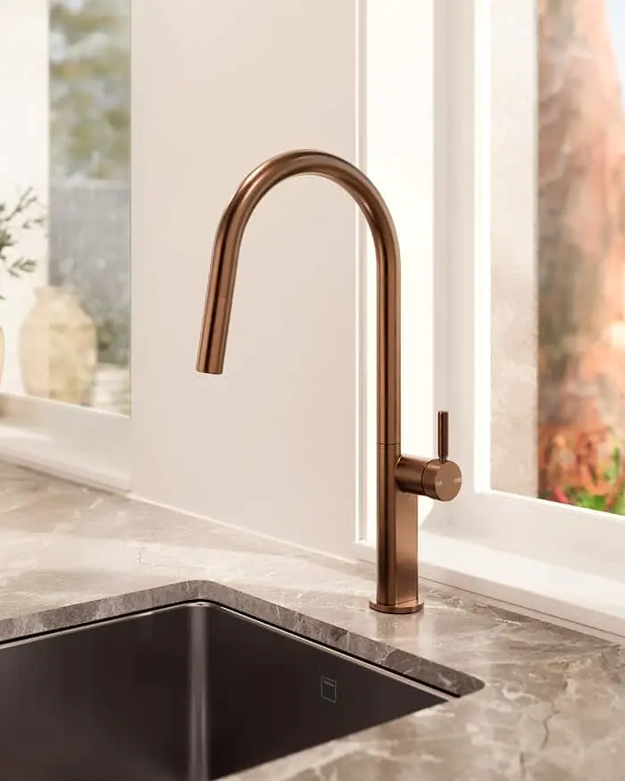 Modern copper kitchen faucet with high arch spout mounted on gray marble countertop next to dark sink by window.