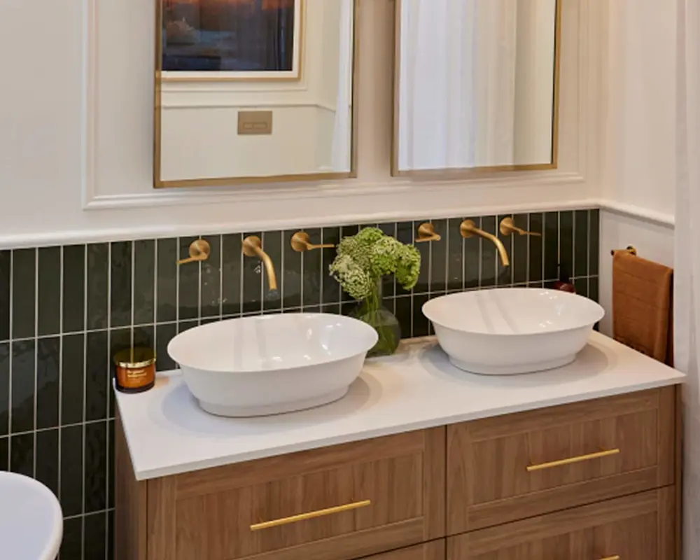 Modern bathroom with dual white vessel sinks on wooden vanity, green tile backsplash, brass faucets, and small plant centerpiece.