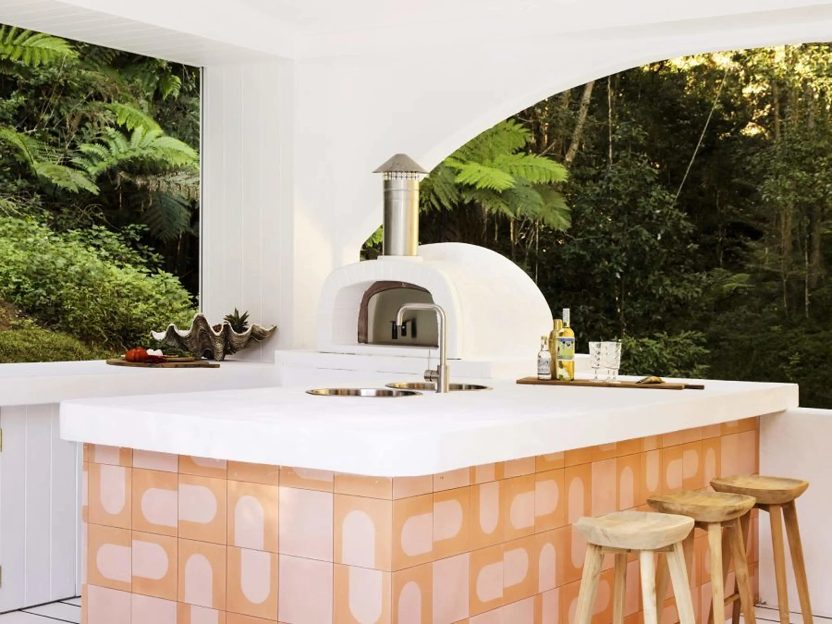 Outdoor kitchen with white pizza oven, peach-tiled counter, wooden stools, and lush forest backdrop with ferns.