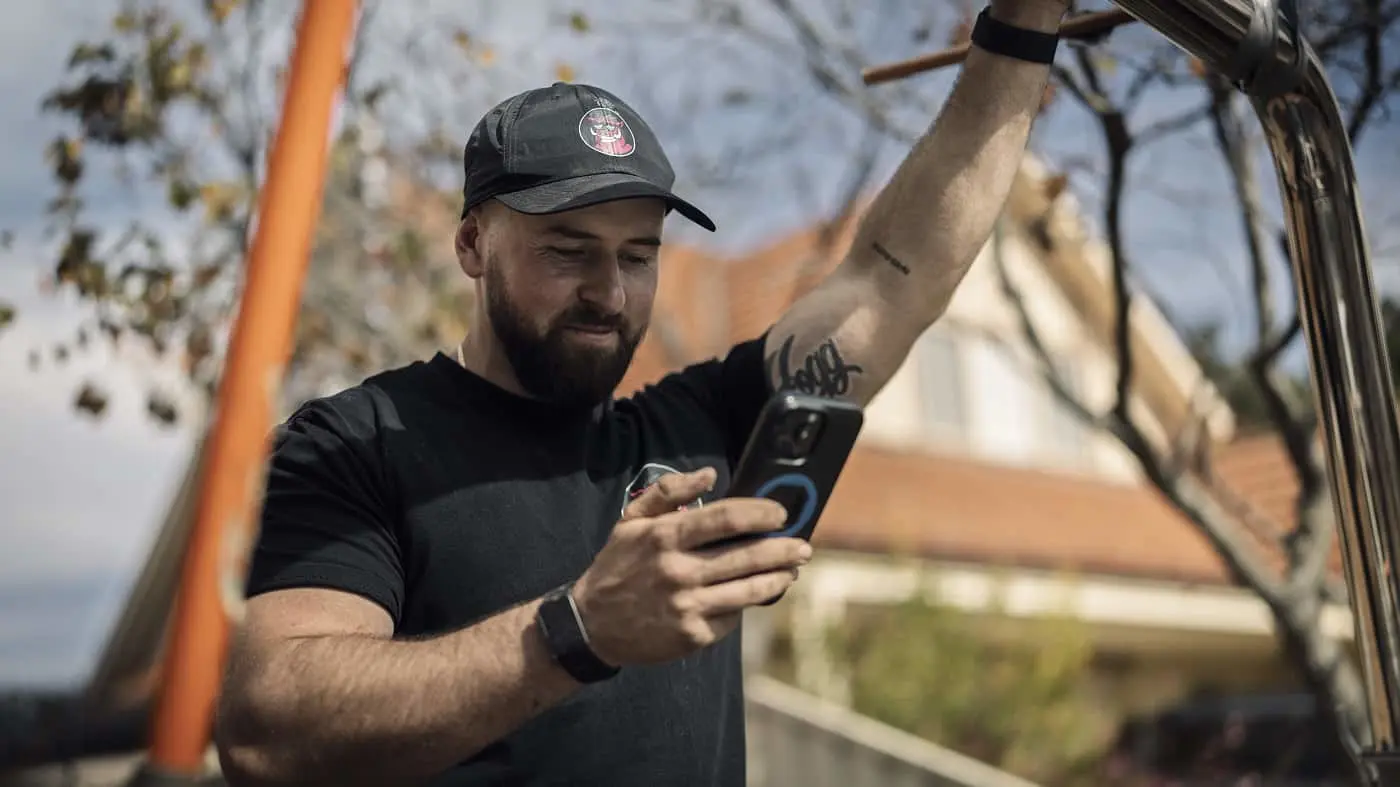 A bearded man wearing a black cap and t-shirt looks at his smartphone outdoors near a vehicle.