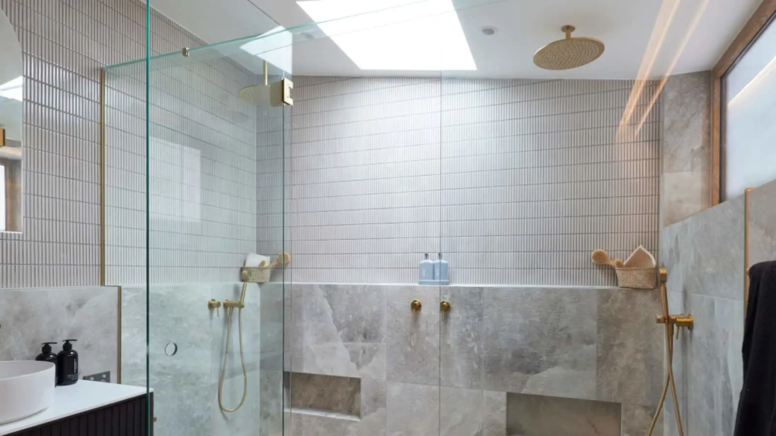 Modern bathroom with glass shower, marble tiles, white vertical wall tiles, brass fixtures, and a skylight above.