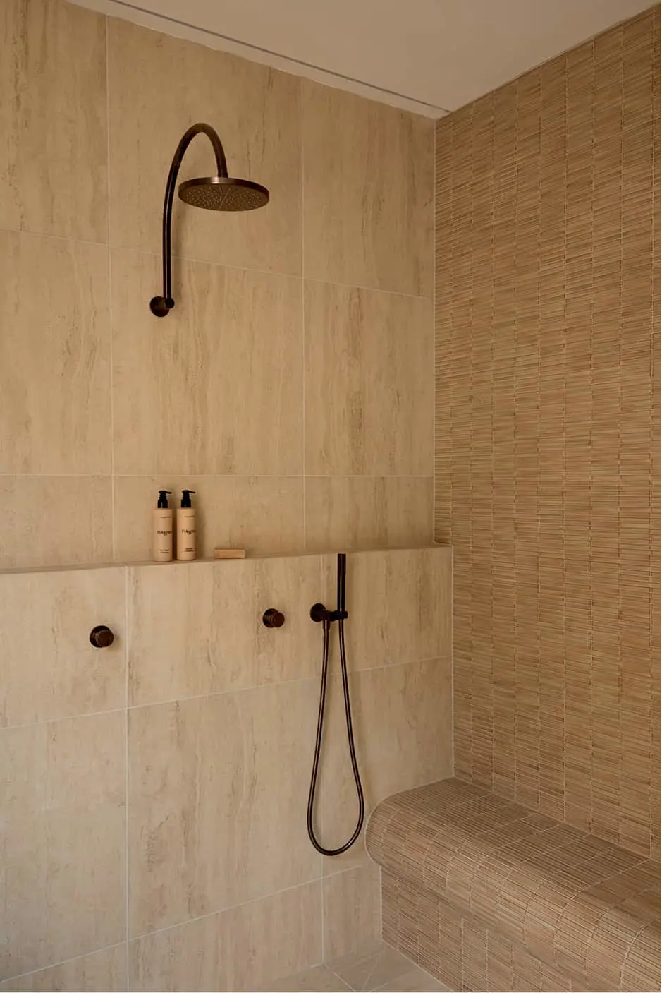 A tiled shower with beige stone walls, a bronze rain showerhead, handheld shower, built-in shelf with toiletries, and a corner bench.