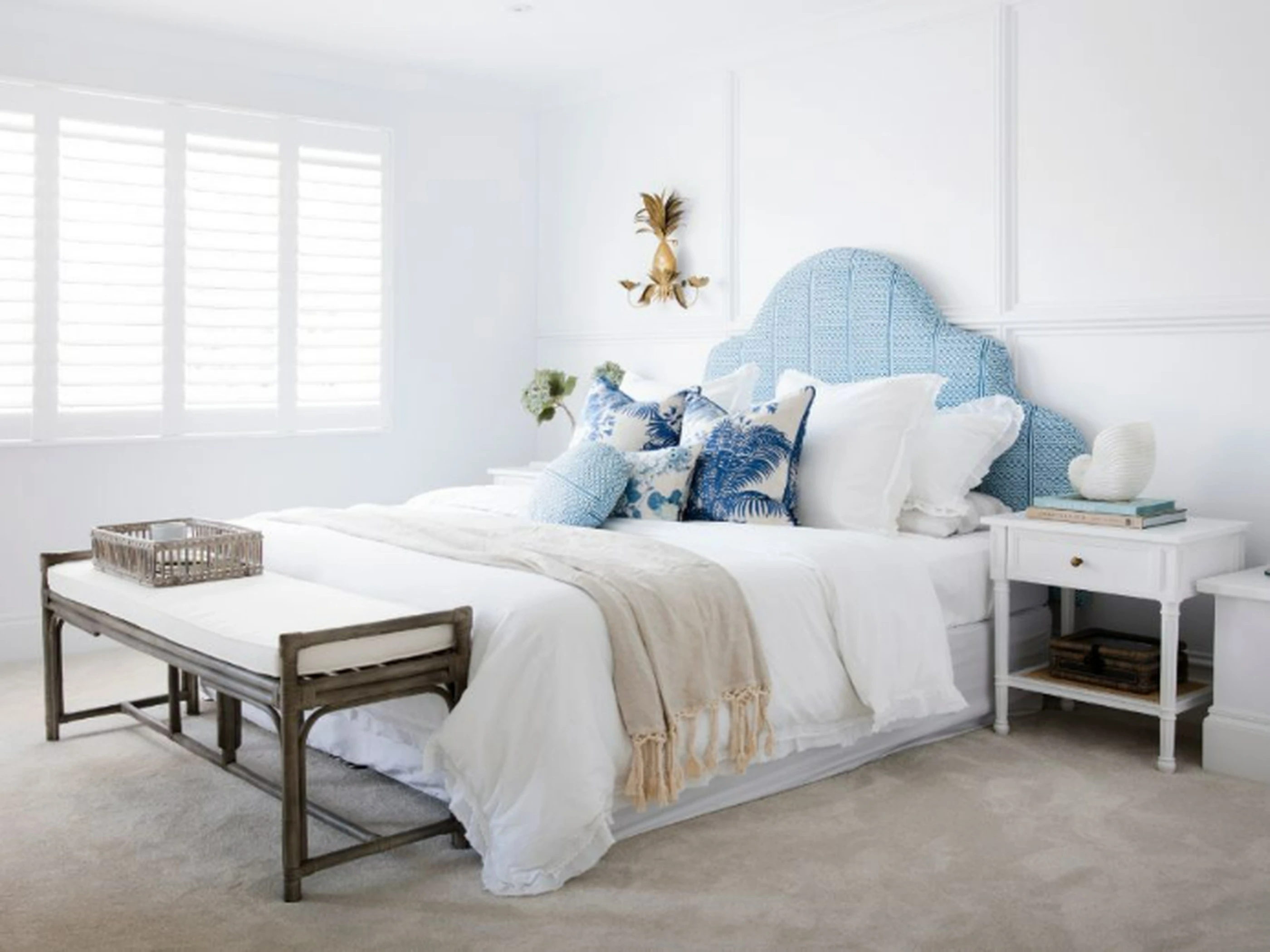 Bright coastal bedroom with blue upholstered headboard, white bedding, blue accent pillows, and gold pineapple wall sconce.