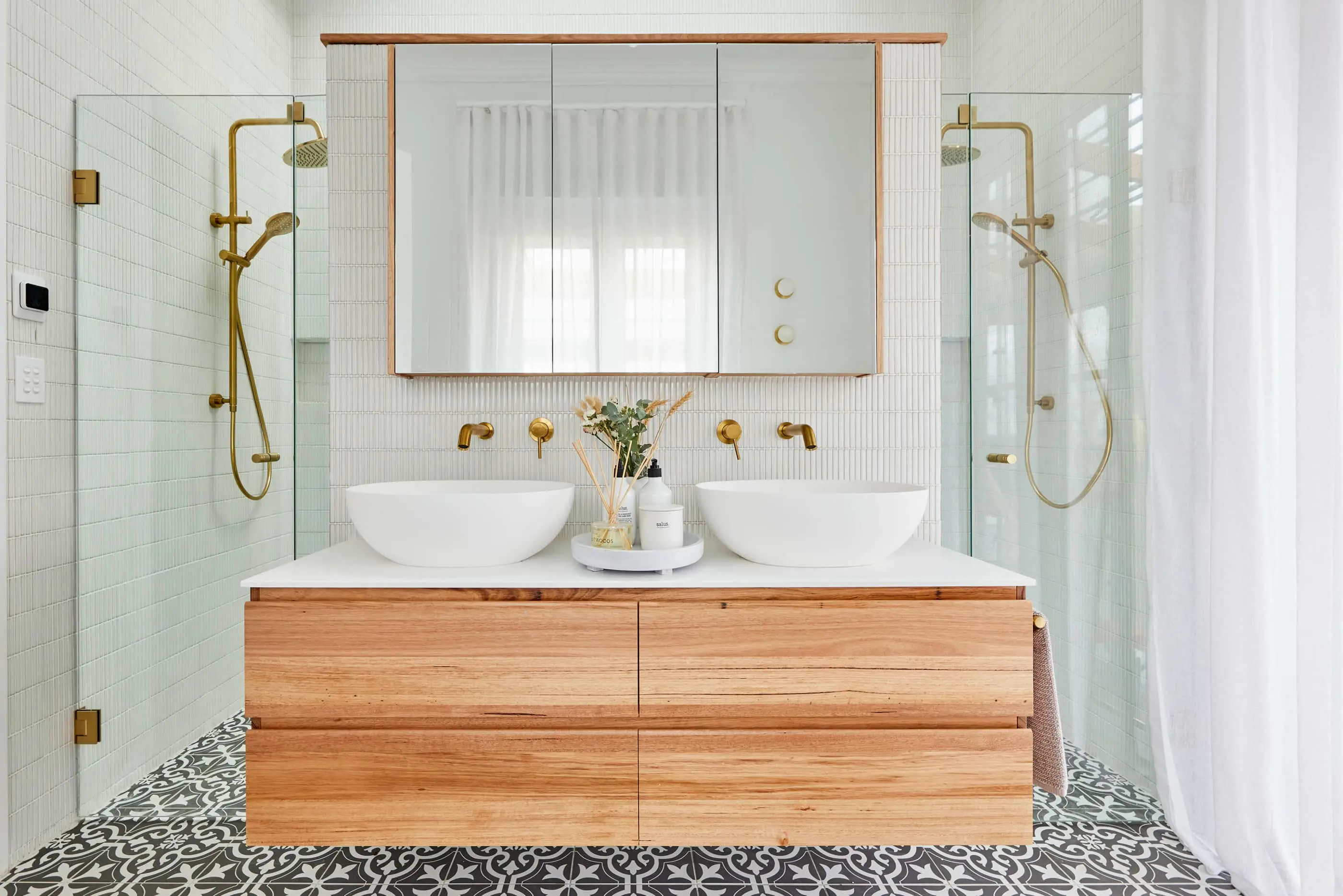 Modern bathroom with white tile walls, wooden vanity with dual sinks, gold fixtures, and patterned floor tiles.