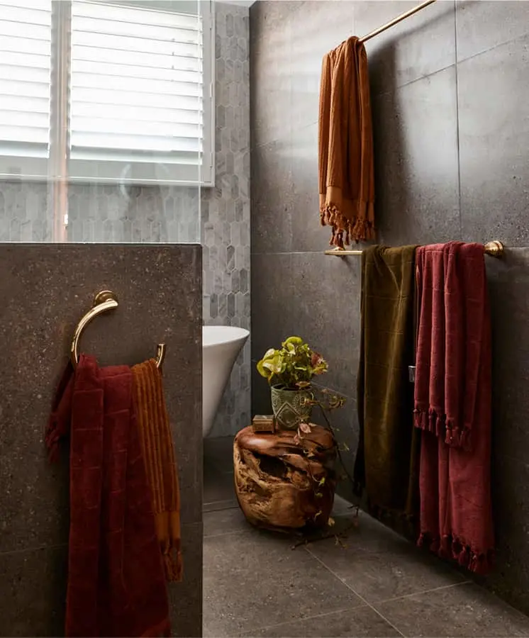 Modern bathroom with gray tile walls, colorful towels in rust and burgundy, wooden stool with small plant, and window.