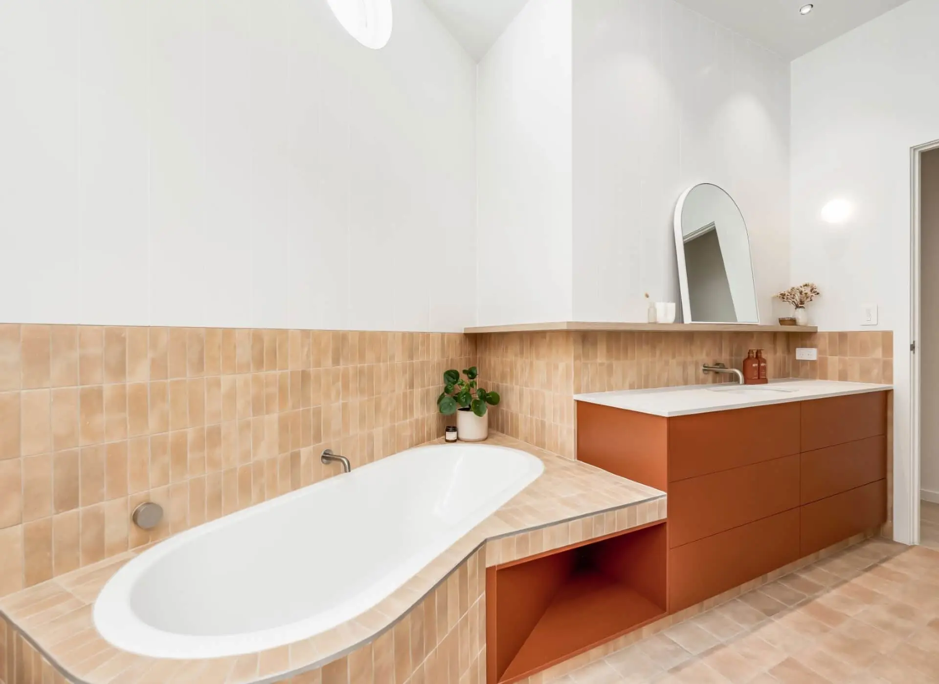 Modern bathroom with white bathtub, terracotta vanity, beige tile backsplash, and minimalist oval mirror on wooden shelf.