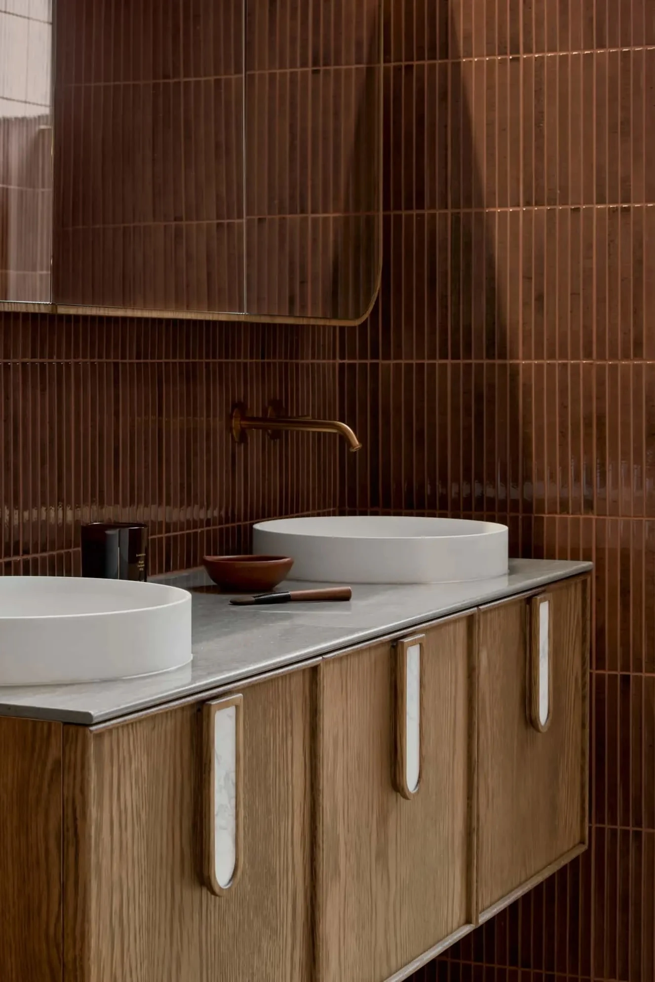 Modern bathroom with brown tiled walls, wooden vanity, white vessel sinks, and brass wall-mounted faucets.