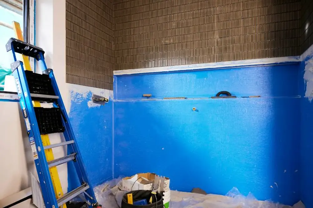 Bathroom renovation in progress with blue waterproofing membrane on walls, brown tiles above, and a ladder nearby.