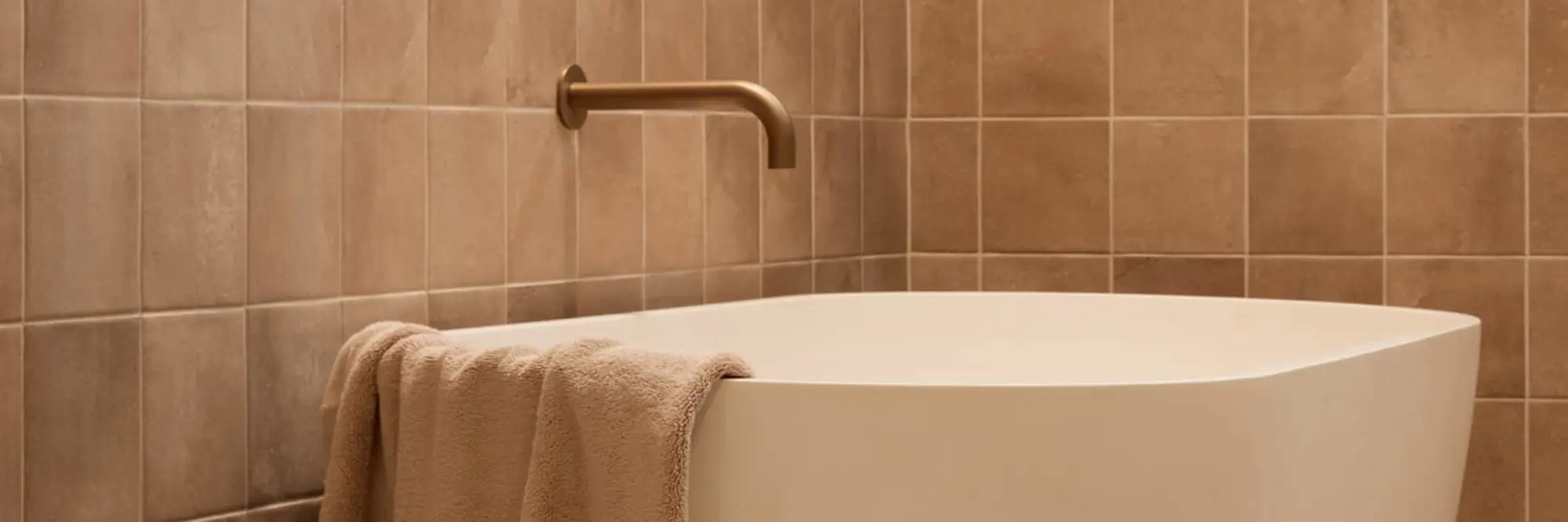 Modern white freestanding bathtub with beige towel against brown tiled wall and brass wall-mounted faucet.
