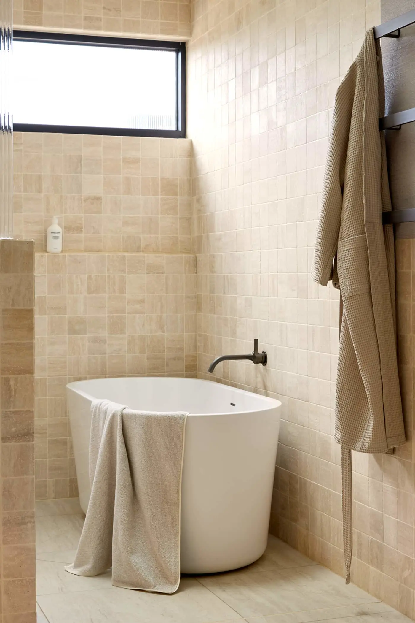 Modern bathroom with white soaking tub, beige tiled walls, hanging towel, and wall-mounted robe on rack.