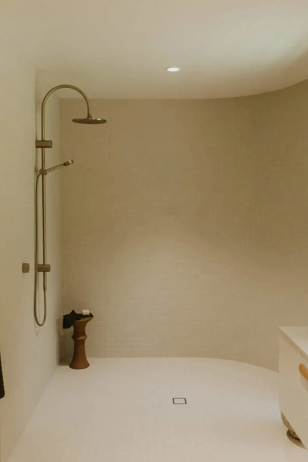 Modern shower with brass rainfall showerhead and handheld attachment in a minimalist beige tiled bathroom with wooden stool.