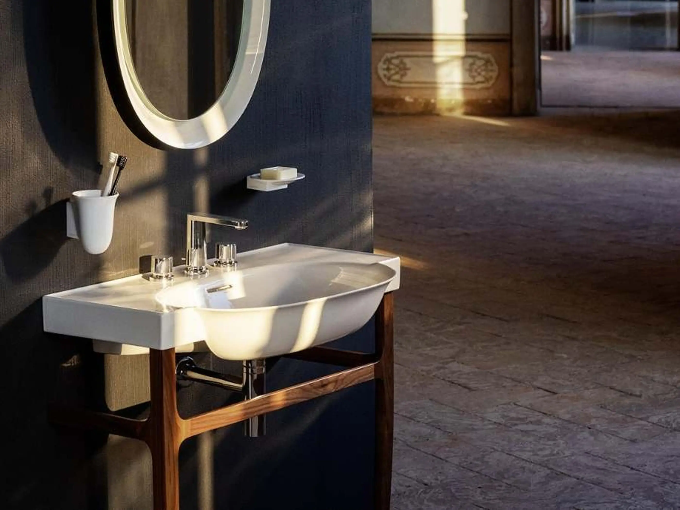 Modern bathroom with white sink on wooden stand, illuminated round mirror, and stone floor in warm lighting.