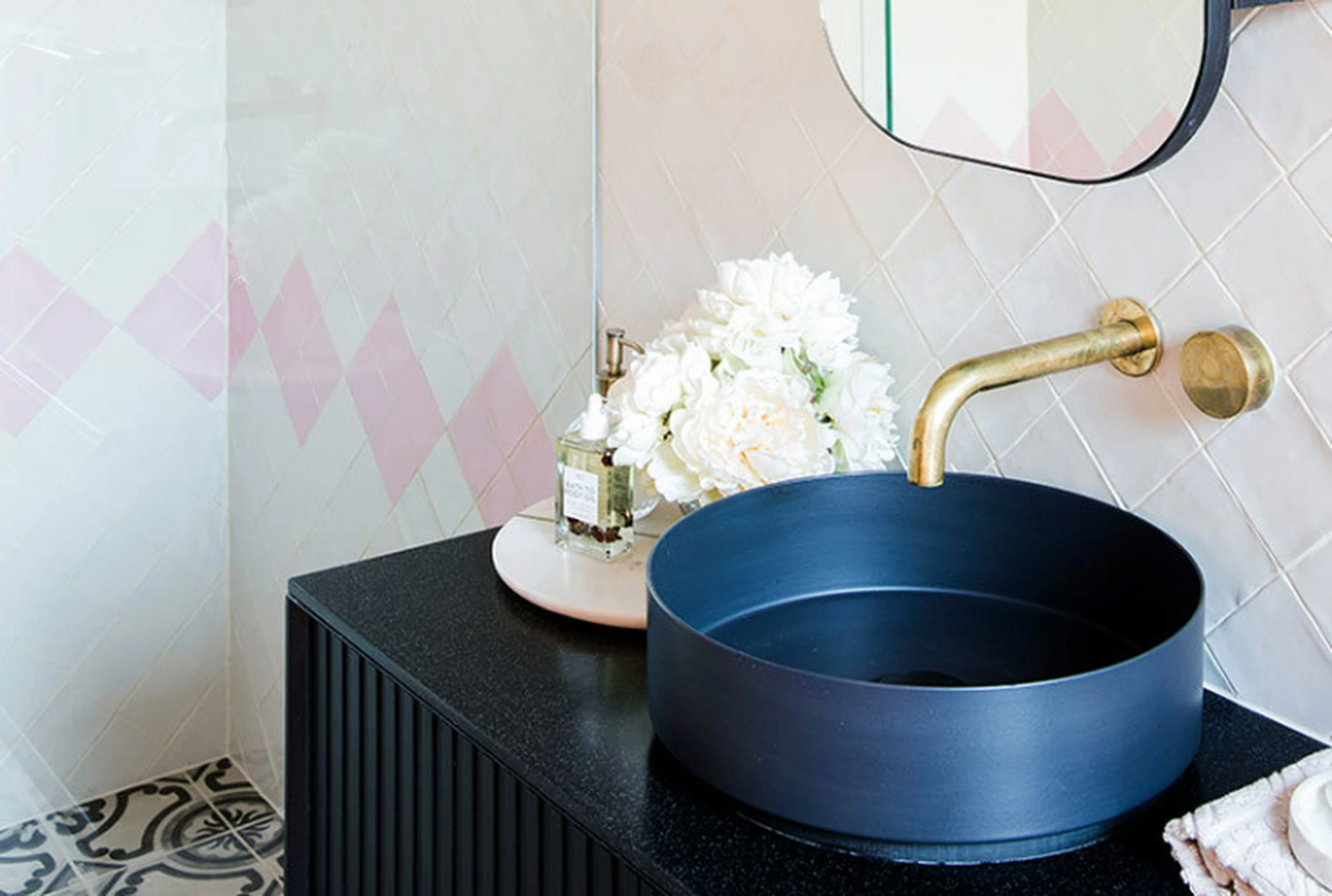 Modern bathroom with navy blue vessel sink, brass faucet, white flowers, and pink diamond-patterned tile wall.