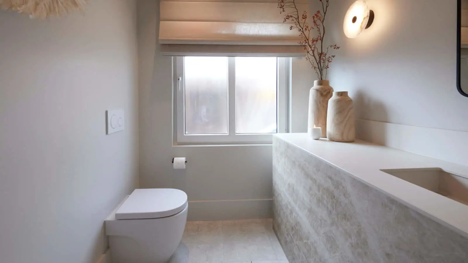 Minimalist bathroom with white toilet, marble countertop, decorative vases, window with roman shade, and wall sconce.