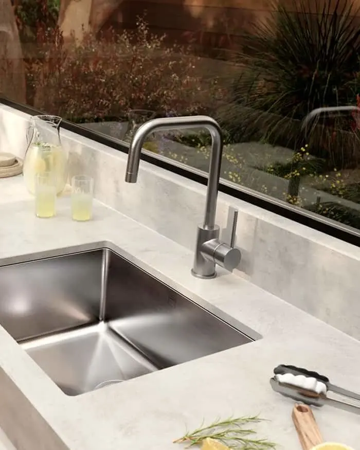 Modern kitchen sink with stainless steel faucet on white marble countertop, garden view through window behind.