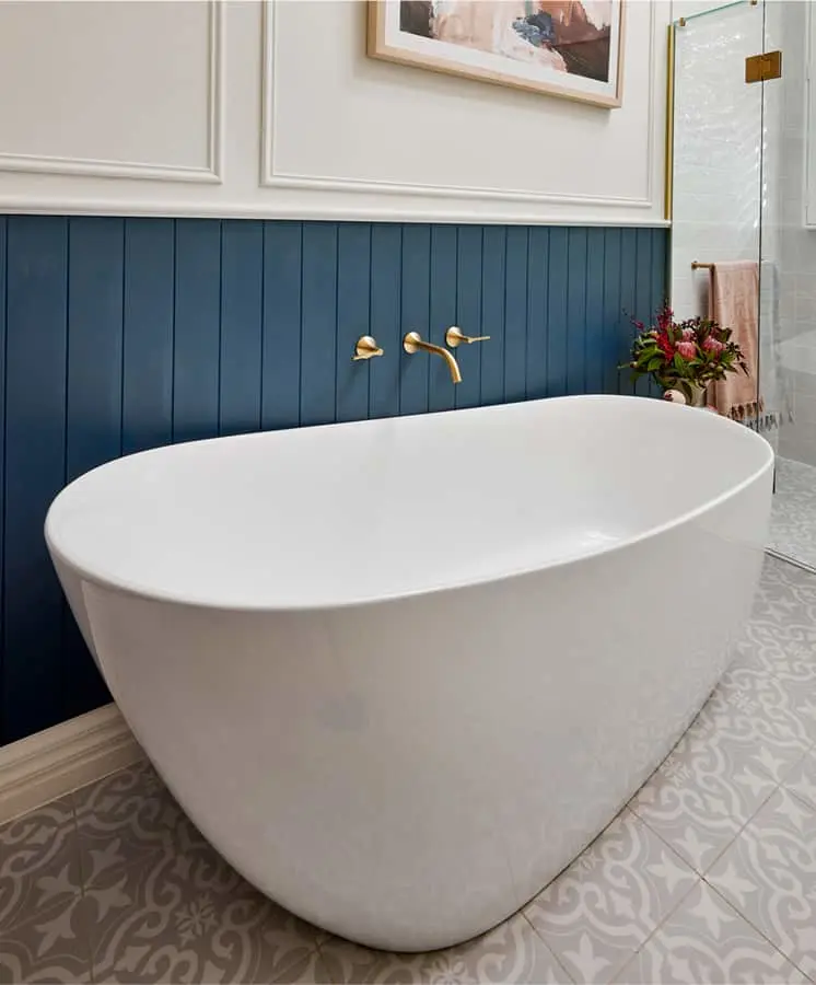 Modern white freestanding bathtub against blue paneled wall with brass wall-mounted faucet and decorative patterned floor tiles.