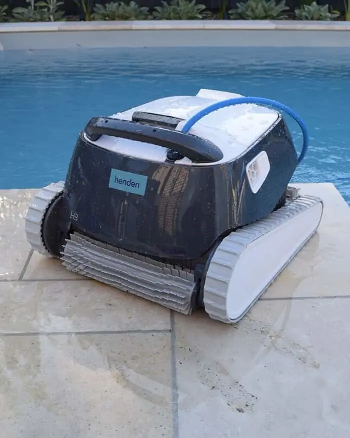 Automatic pool cleaner robot with treads sitting on the edge of a blue swimming pool surrounded by plants.