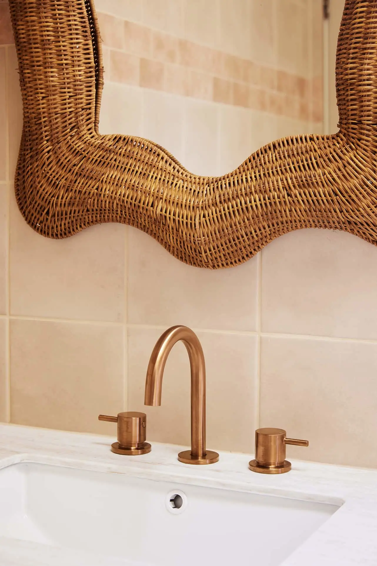 Bathroom sink with copper faucet and woven rattan mirror frame against beige tiled wall.