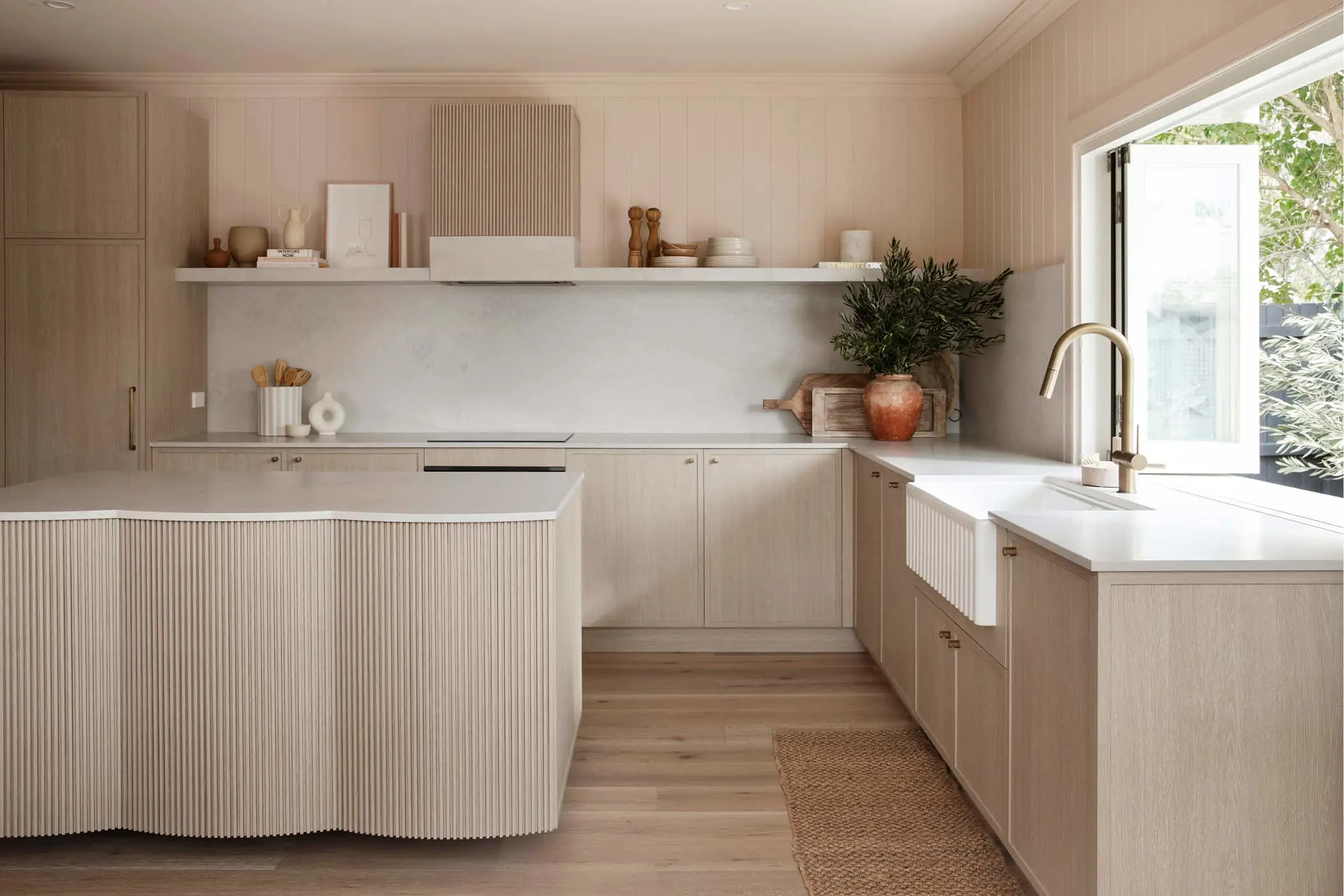 Modern minimalist kitchen with light wood cabinets, white countertops, textured island, and a farmhouse sink near a window.