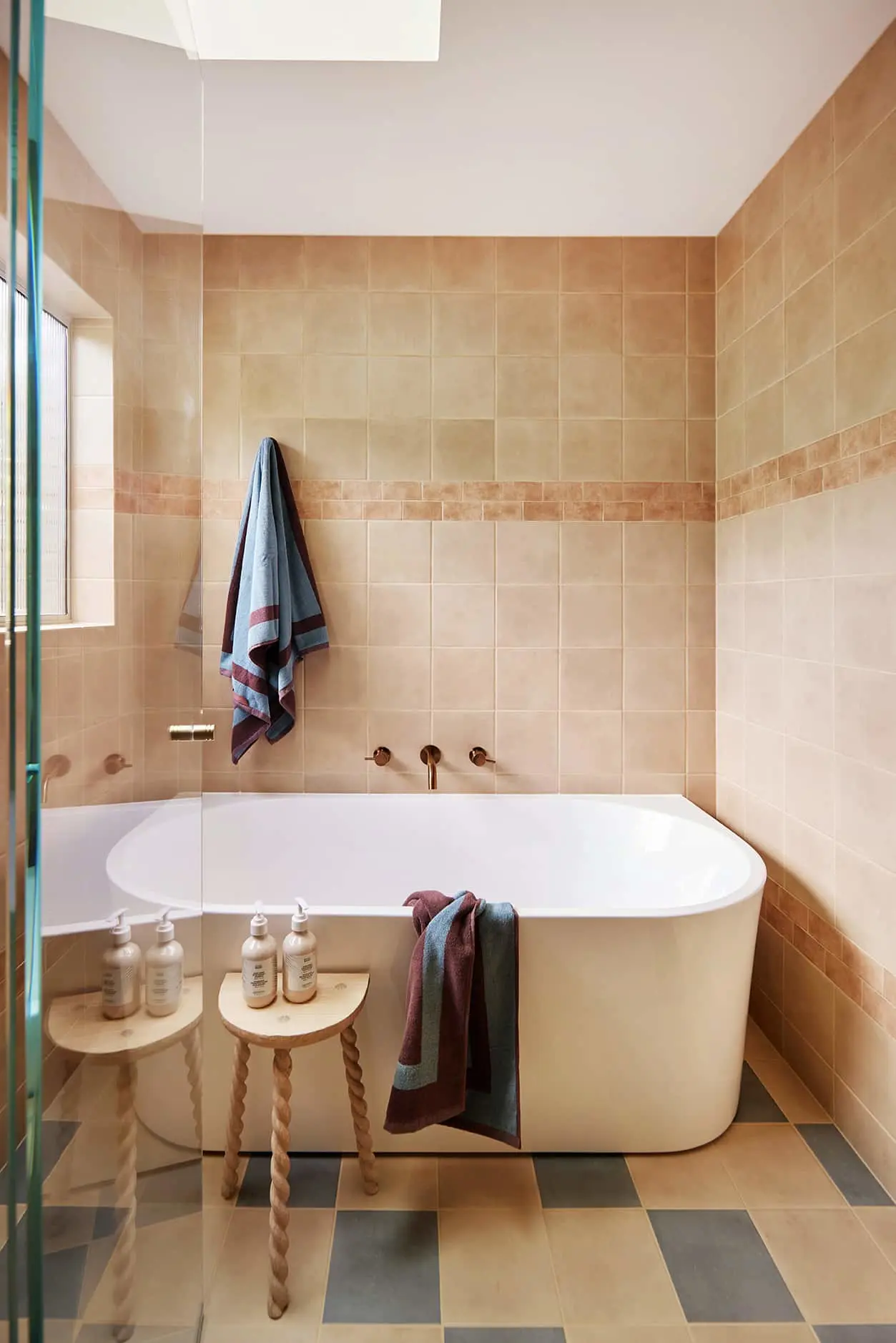 Modern bathroom with beige tiled walls, white freestanding tub, wooden side table with toiletries, and blue-burgundy towels.