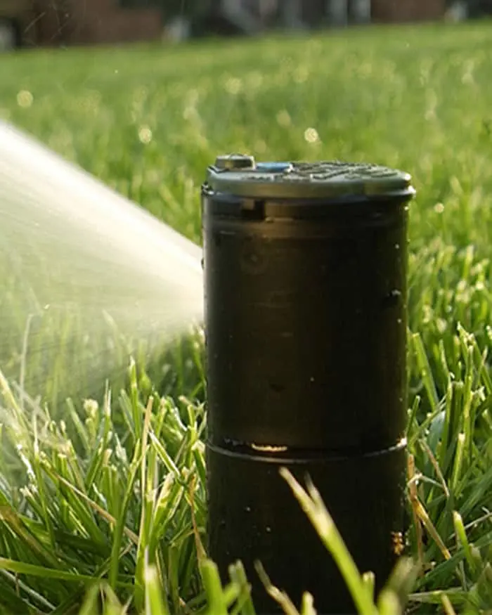 Black lawn sprinkler head in green grass spraying water from the side, sunlight illuminating the water spray.