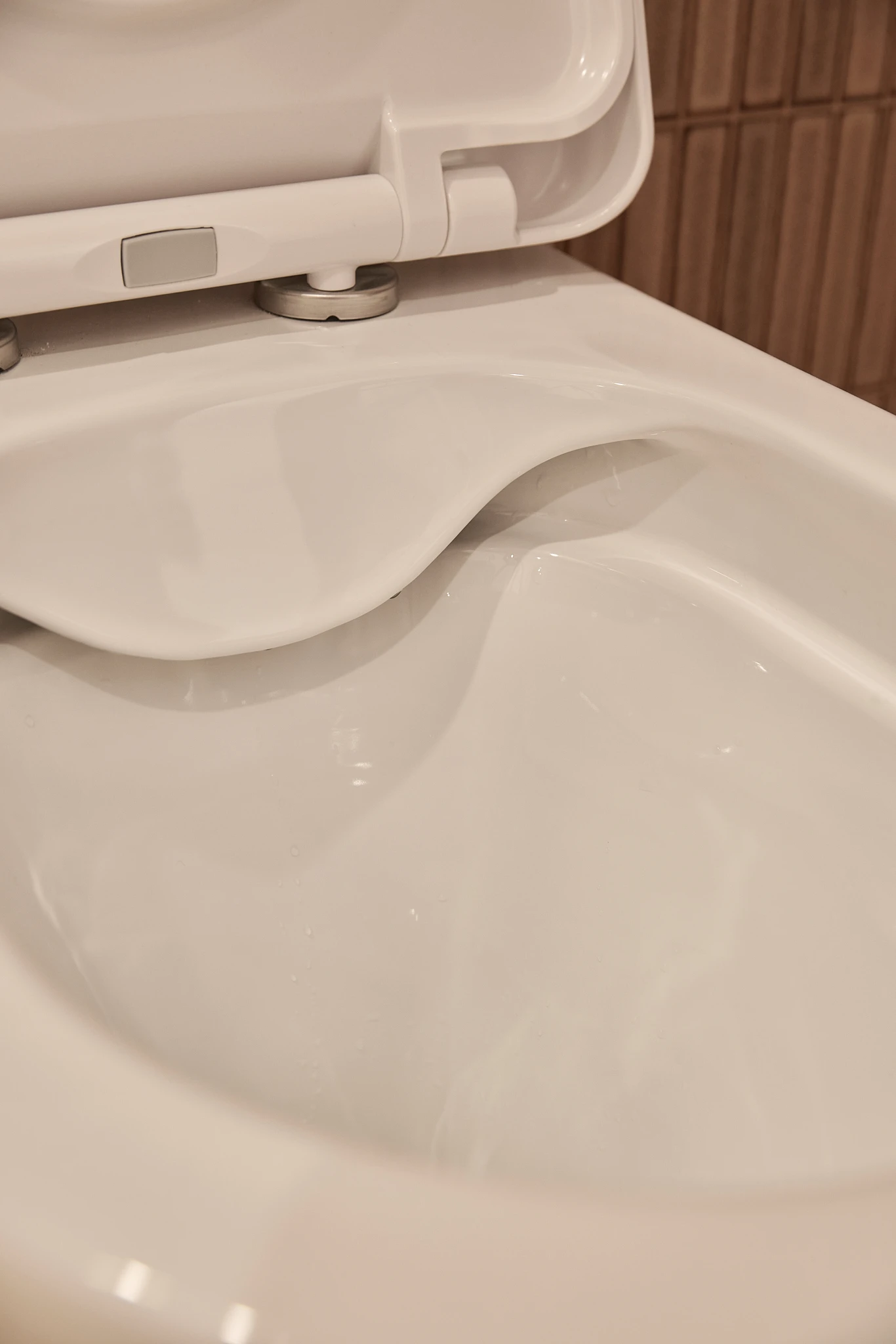 Close-up of a white toilet bowl interior showing the rim and seat hinge against a brown tiled bathroom wall.