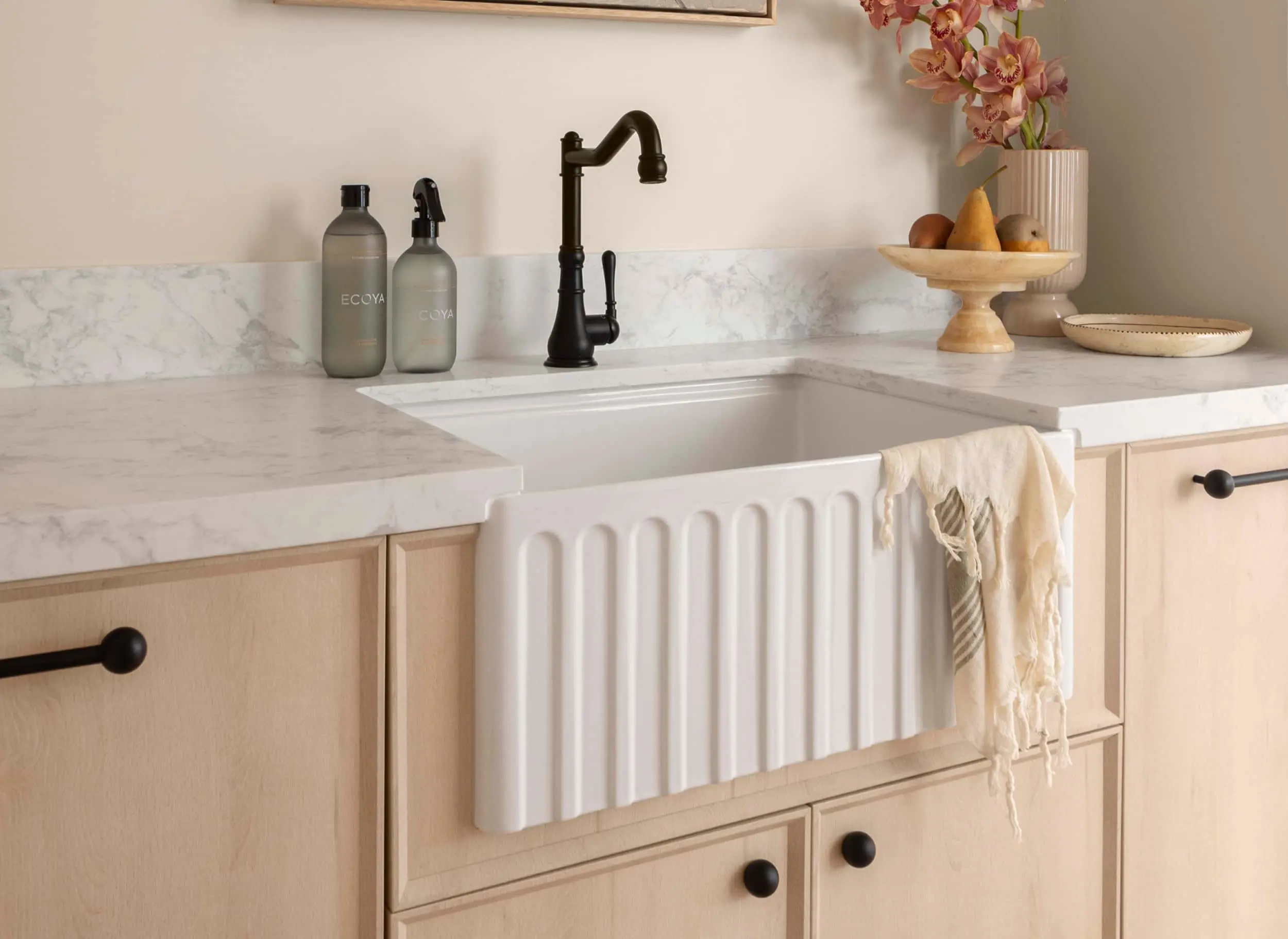 Elegant kitchen with white farmhouse sink, black faucet, marble countertops, wooden cabinets, and decorative fruit bowl.
