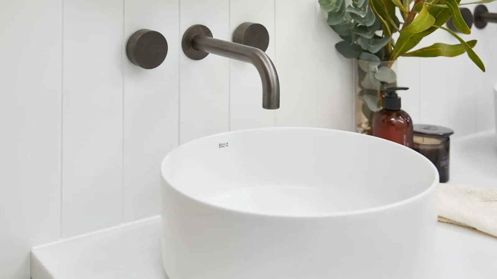 Modern white vessel sink with wall-mounted bronze faucet, decorative plants and amber bottle on counter.