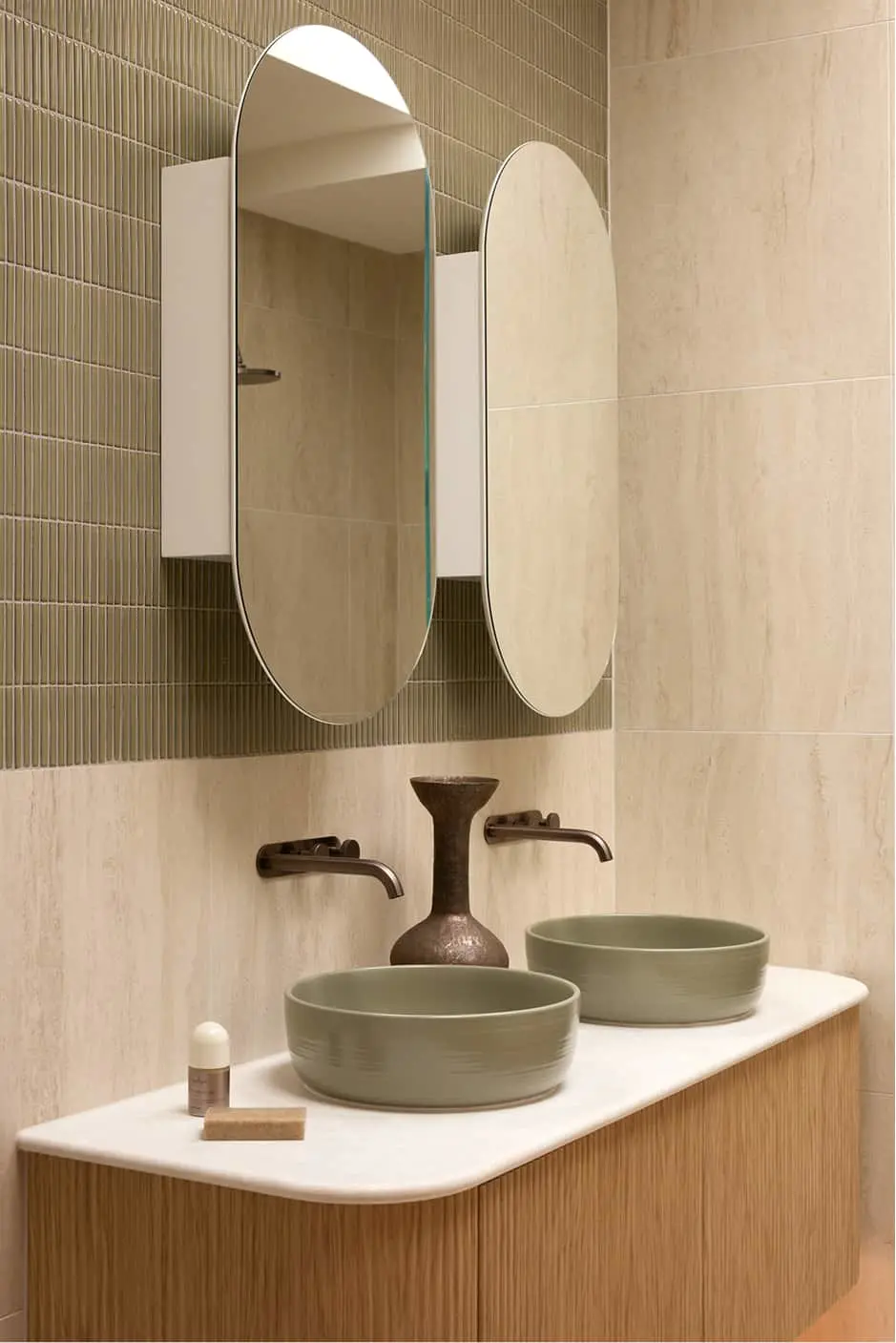 Modern bathroom with two oval mirrors above green vessel sinks on white countertop with wood cabinet and beige tile walls.