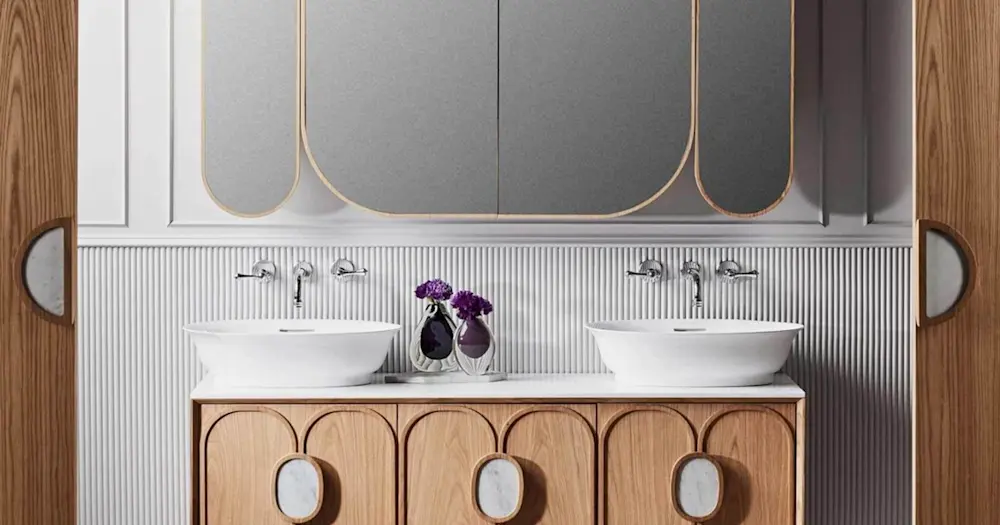 Modern bathroom with double white vessel sinks, wooden vanity, gold-trimmed mirrors, and purple flowers in vases.