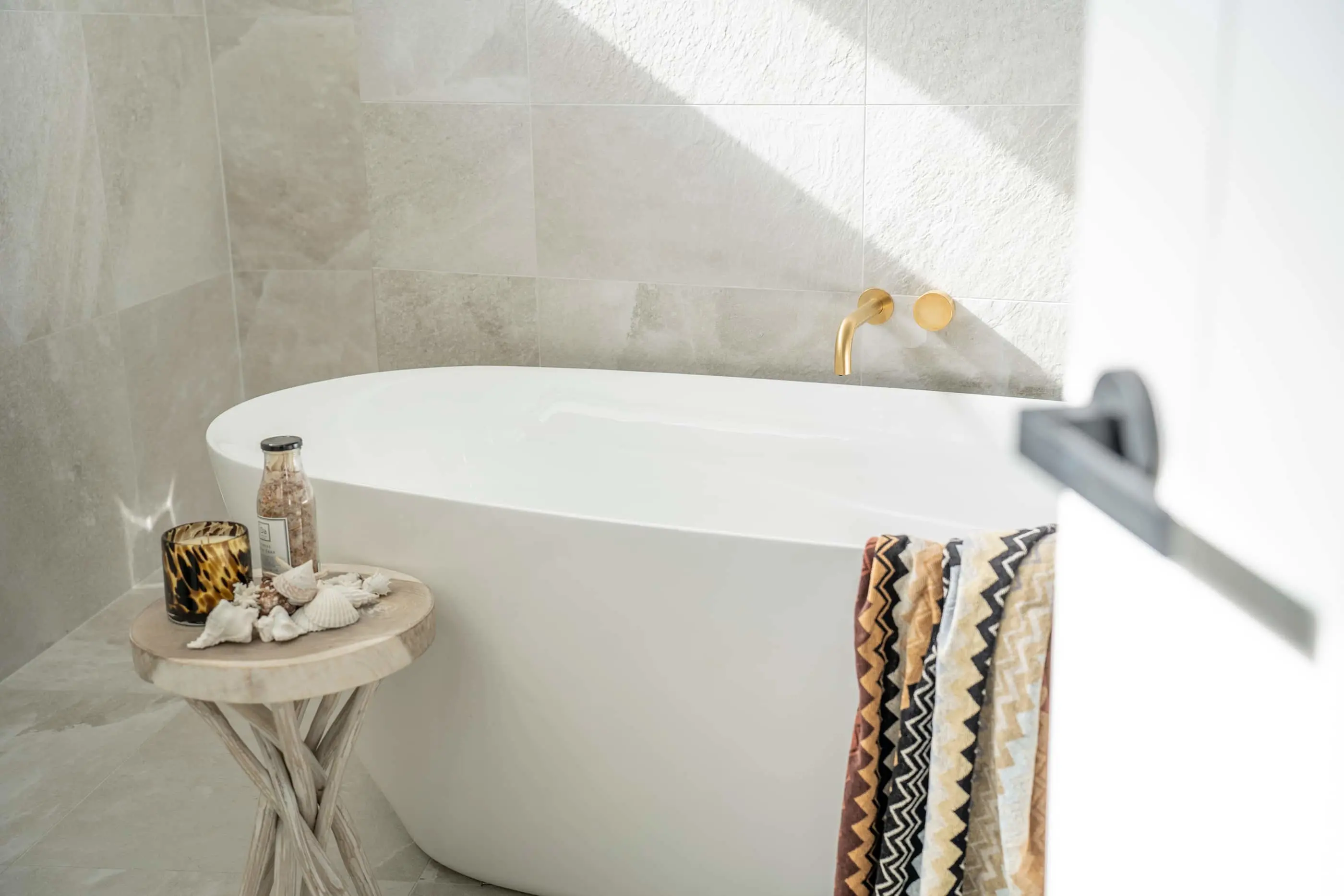 Modern white bathtub with gold fixtures, patterned towel, and small side table holding bath salts and decorative items.