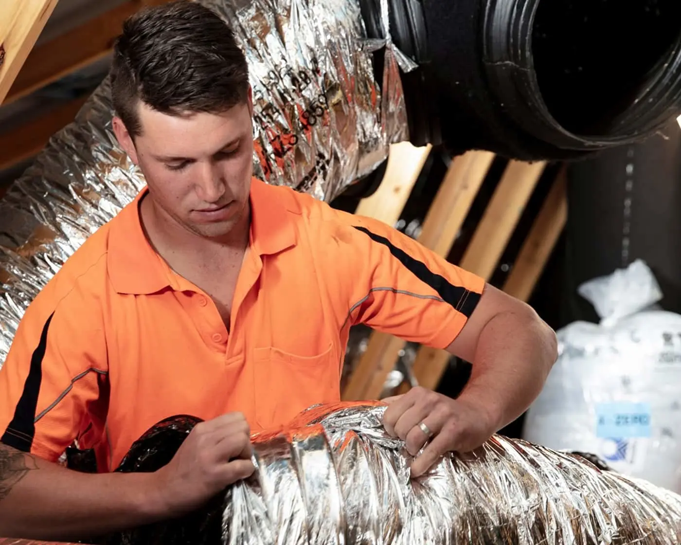Technician in bright orange work shirt installing reflective insulation in an attic or crawl space with ductwork visible.