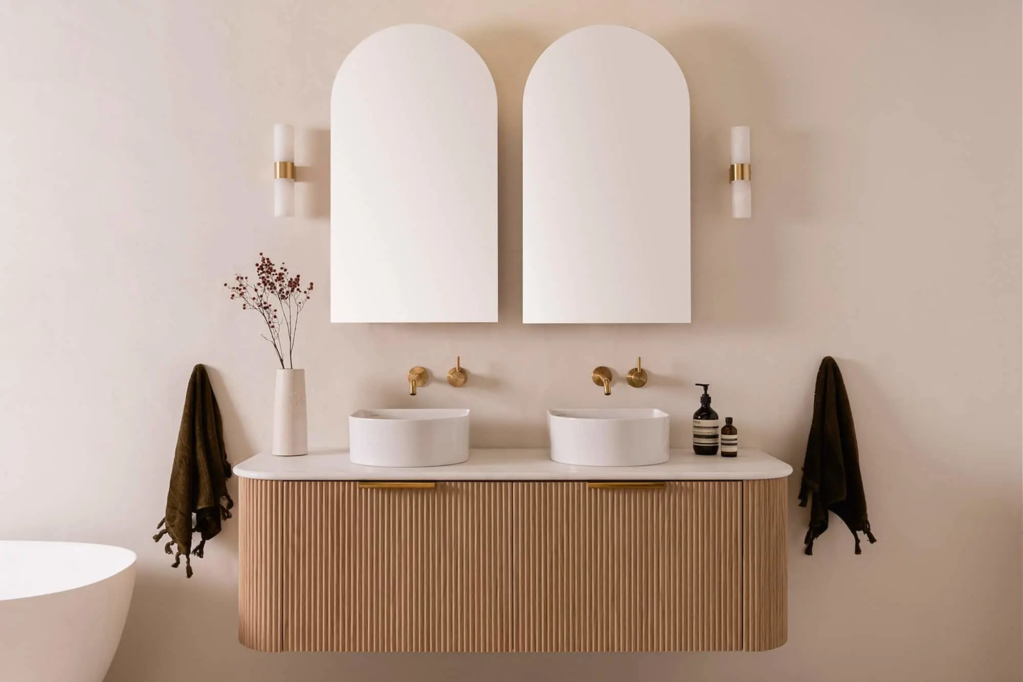 Modern bathroom with dual white sinks on wooden vanity, arched mirrors, brass fixtures, and minimalist decor.