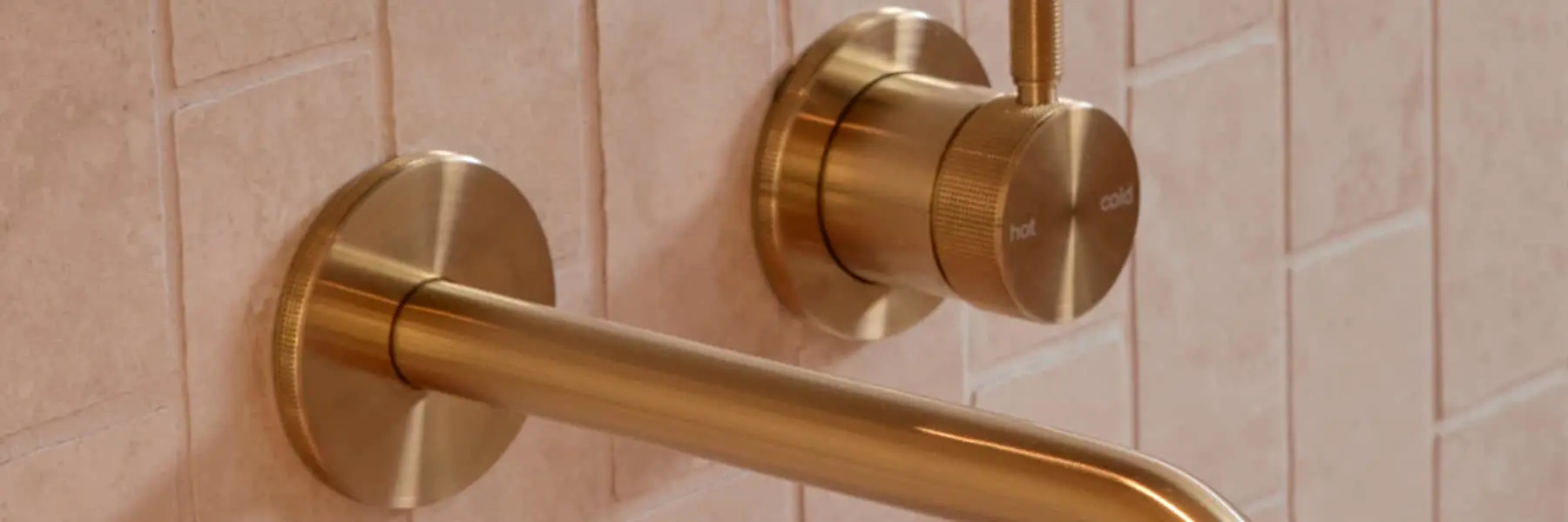 Brass bathroom faucet with wall-mounted spout and temperature control knob against light pink tiled wall.