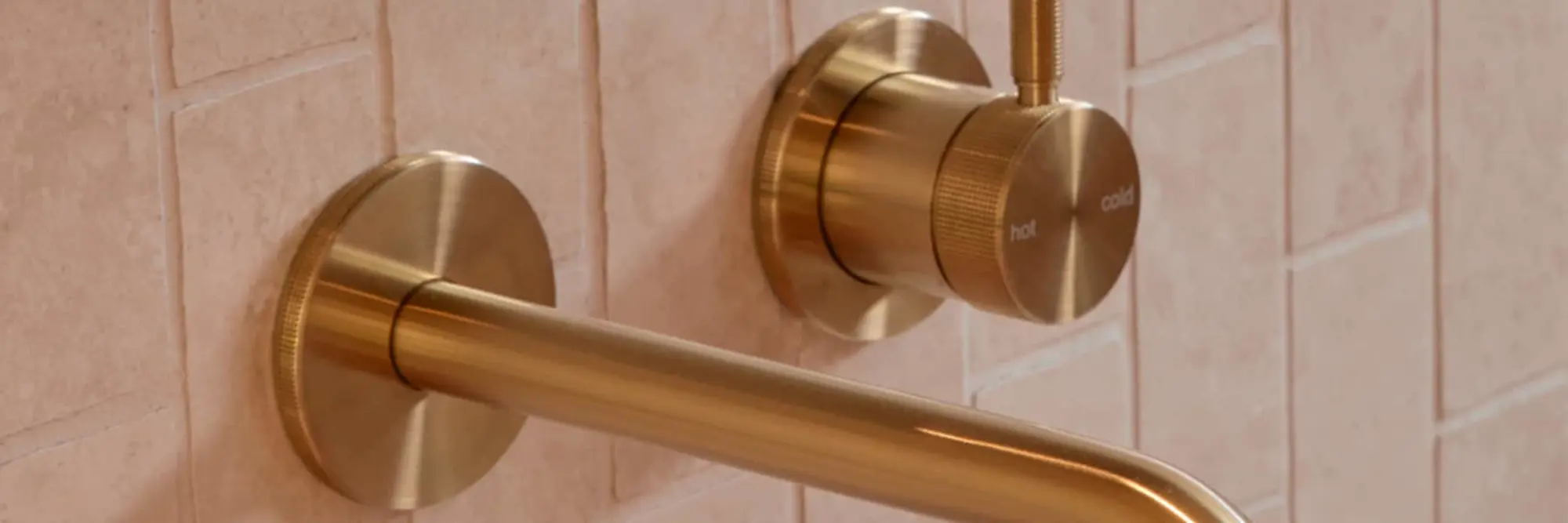 Brass bathroom faucet with wall-mounted spout and temperature control knob against light pink tiled wall.