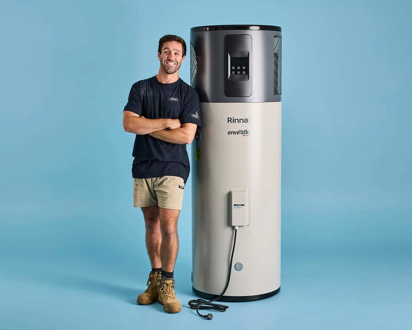 Person in navy shirt and shorts standing next to a Rinnai Enviroflo water heater against blue background.