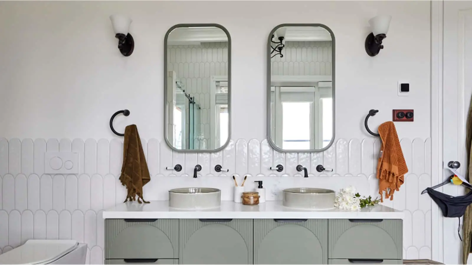 Modern bathroom with double vanity, rounded mirrors, white tile backsplash, and hanging towels in brown and orange.