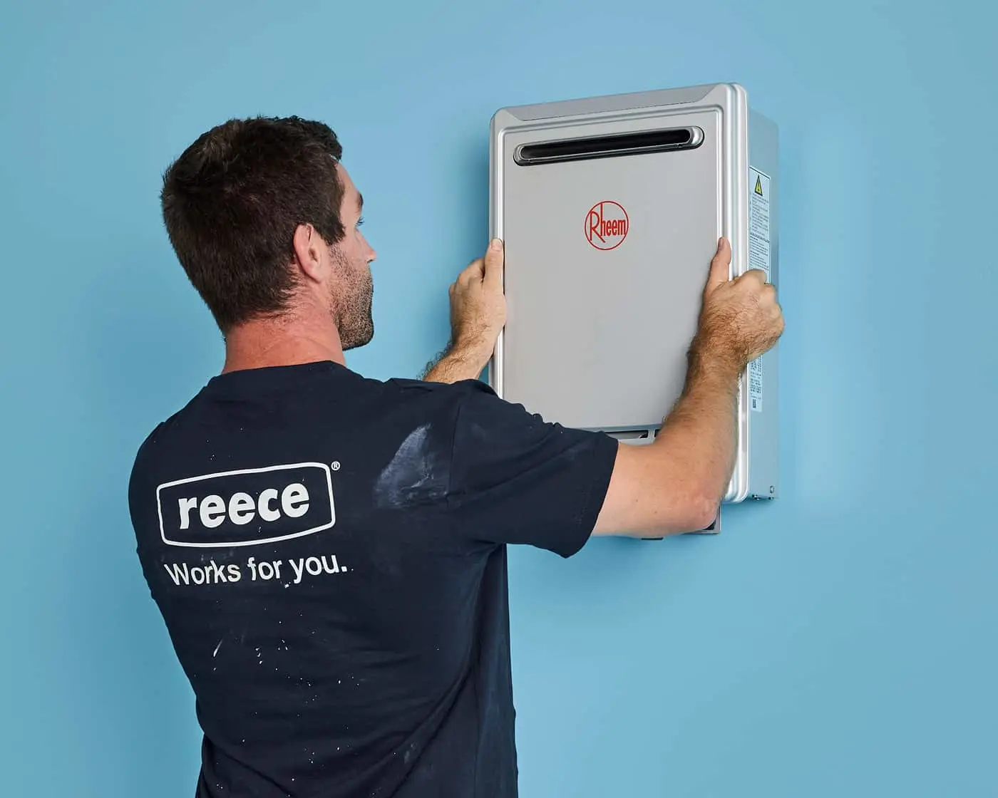 Technician in Reece shirt installing a Rheem water heater unit on a light blue wall.