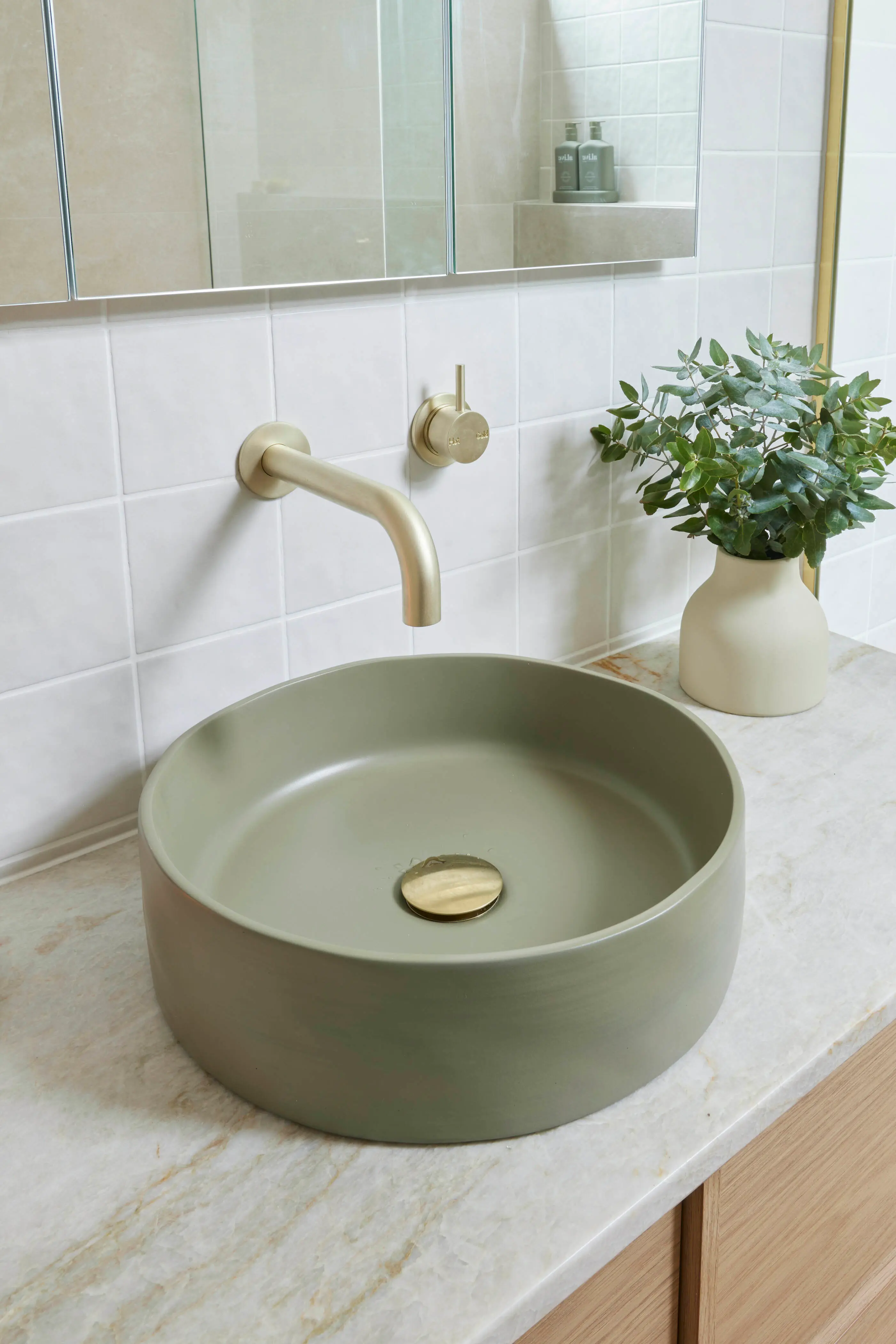 Modern bathroom with sage green vessel sink, brass wall-mounted faucet, white tile backsplash, and small plant in vase.