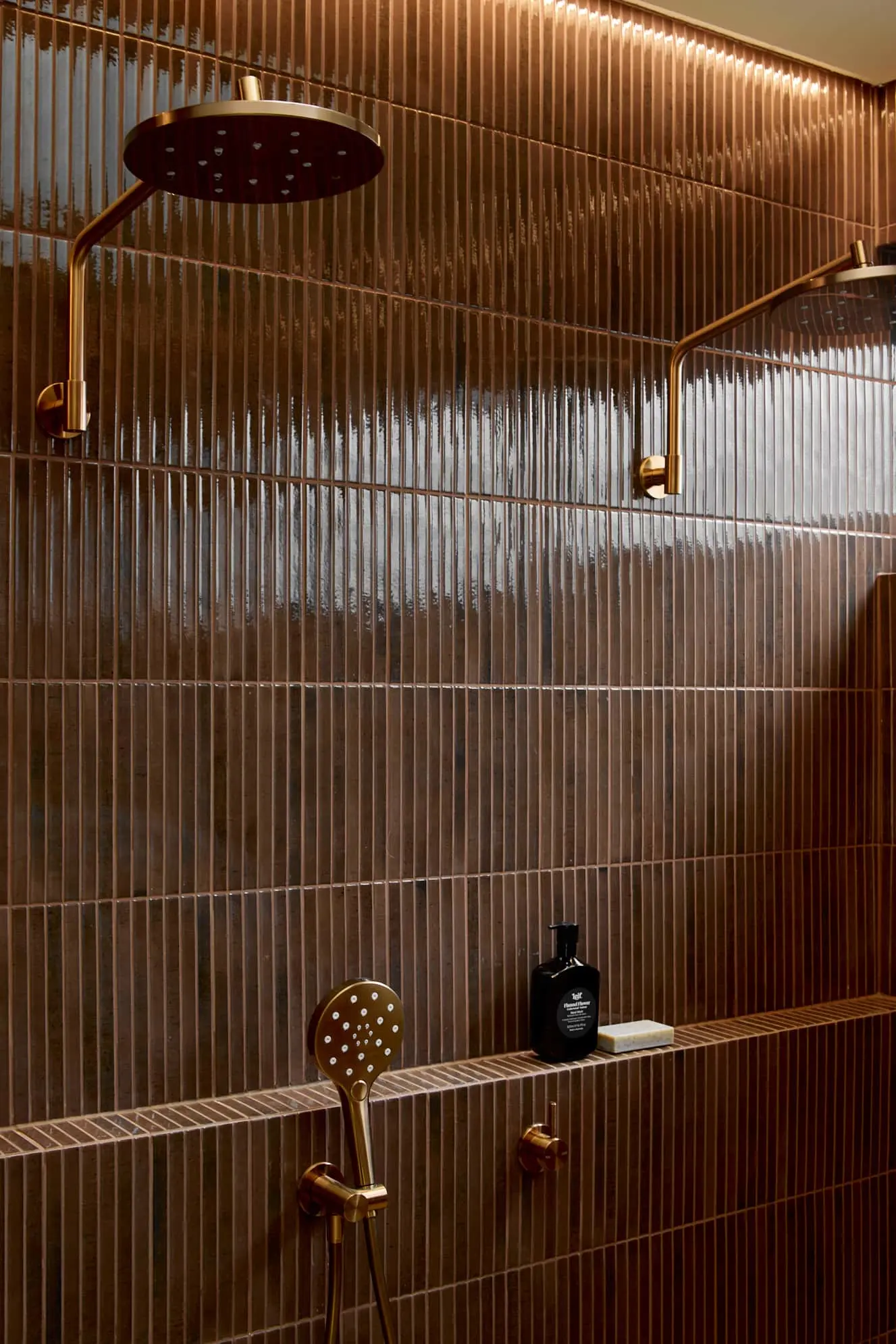 Modern shower with bronze fixtures and rainfall showerhead against vertical brown tiled walls with built-in shelf.