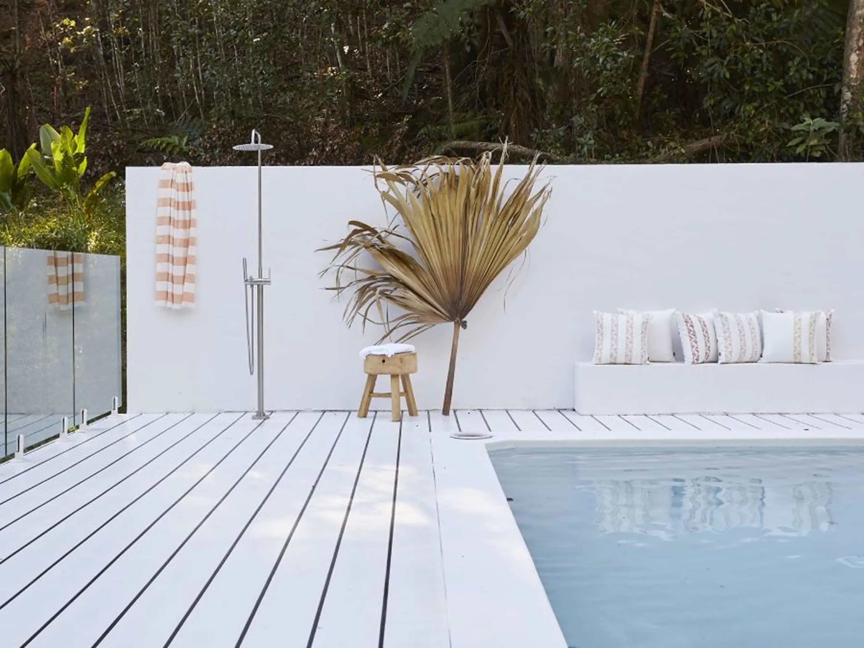 Modern outdoor pool area with white deck, dried palm frond, outdoor shower, and cushioned seating against forest backdrop.