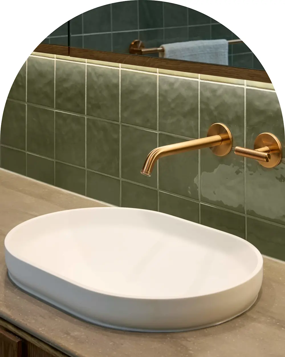 Modern bathroom sink with white oval basin, brass wall-mounted faucet, and green tiled wall with wooden accent.