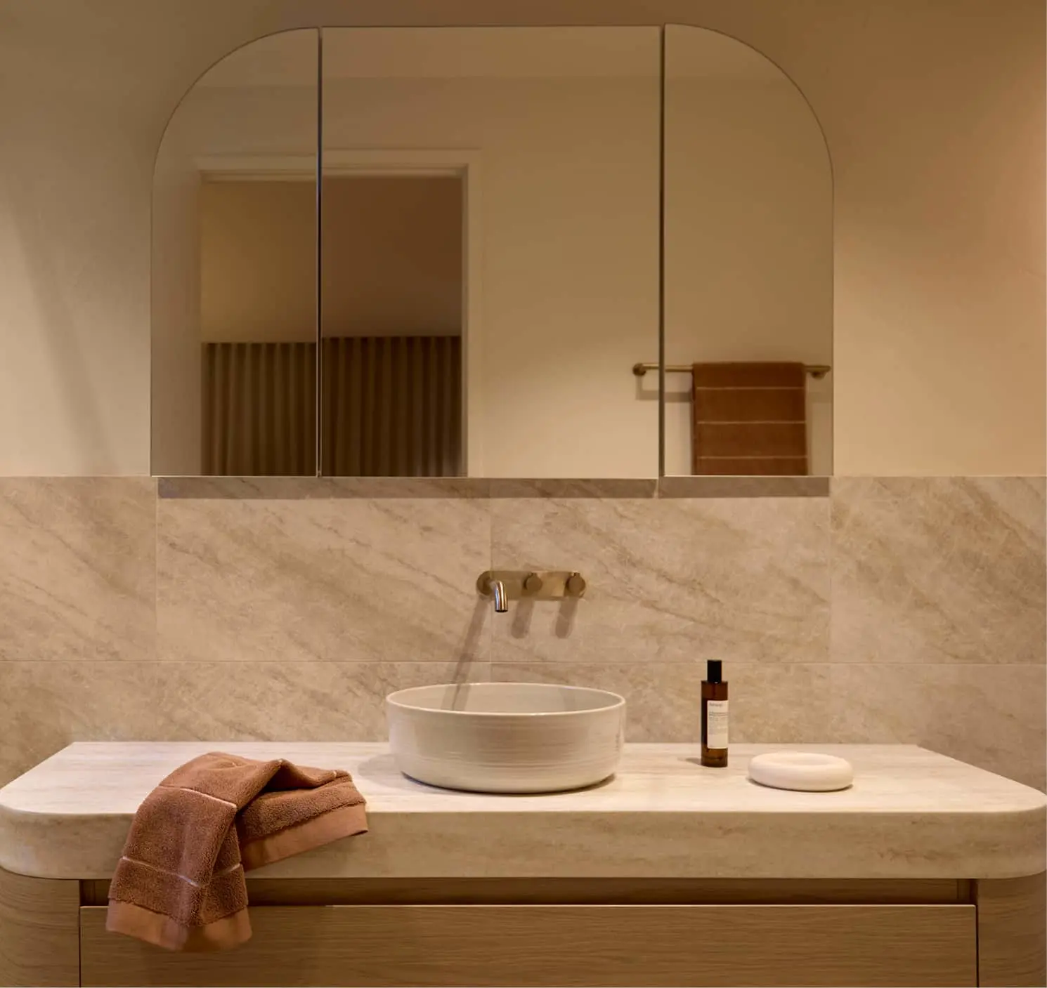 Modern bathroom with arched mirror above white countertop, round sink, brass fixtures, and beige stone walls.