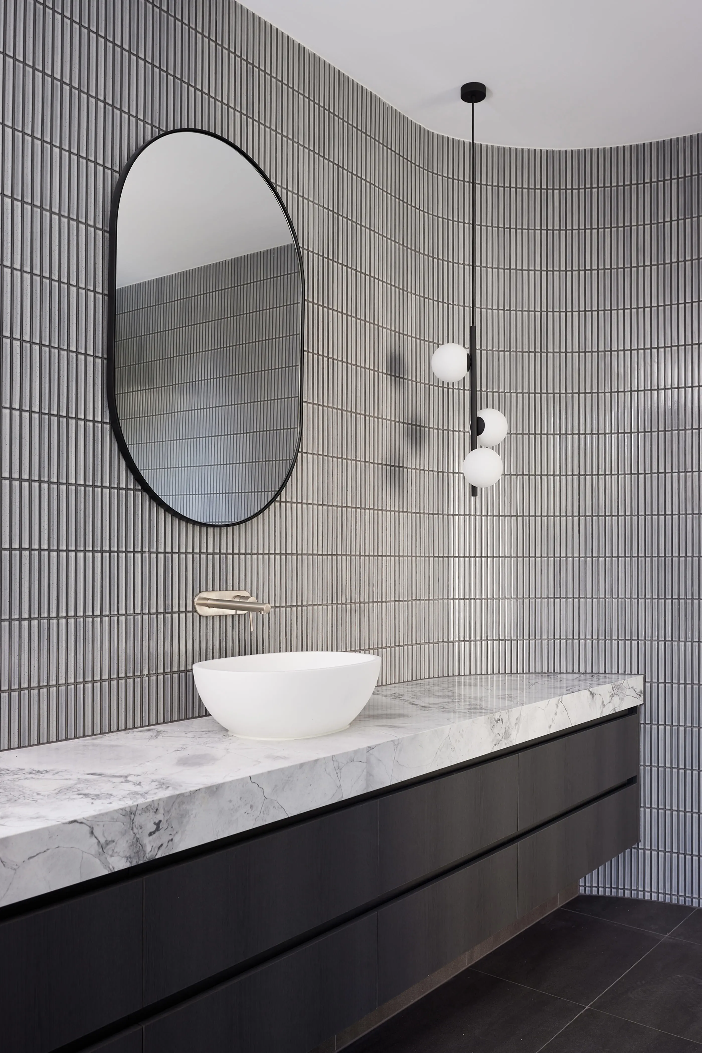 Modern bathroom with gray tiled walls, oval mirror, white vessel sink on marble countertop, and pendant lighting.