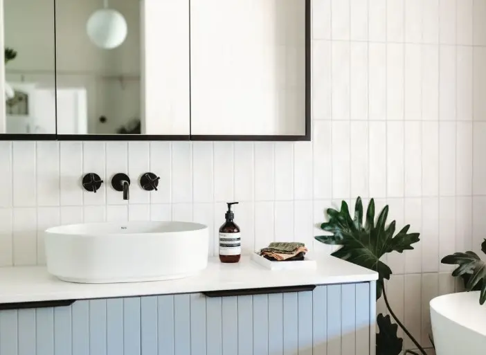 Modern bathroom with white oval sink, light blue vanity, black fixtures, mirror, and green plant on white tiled wall.
