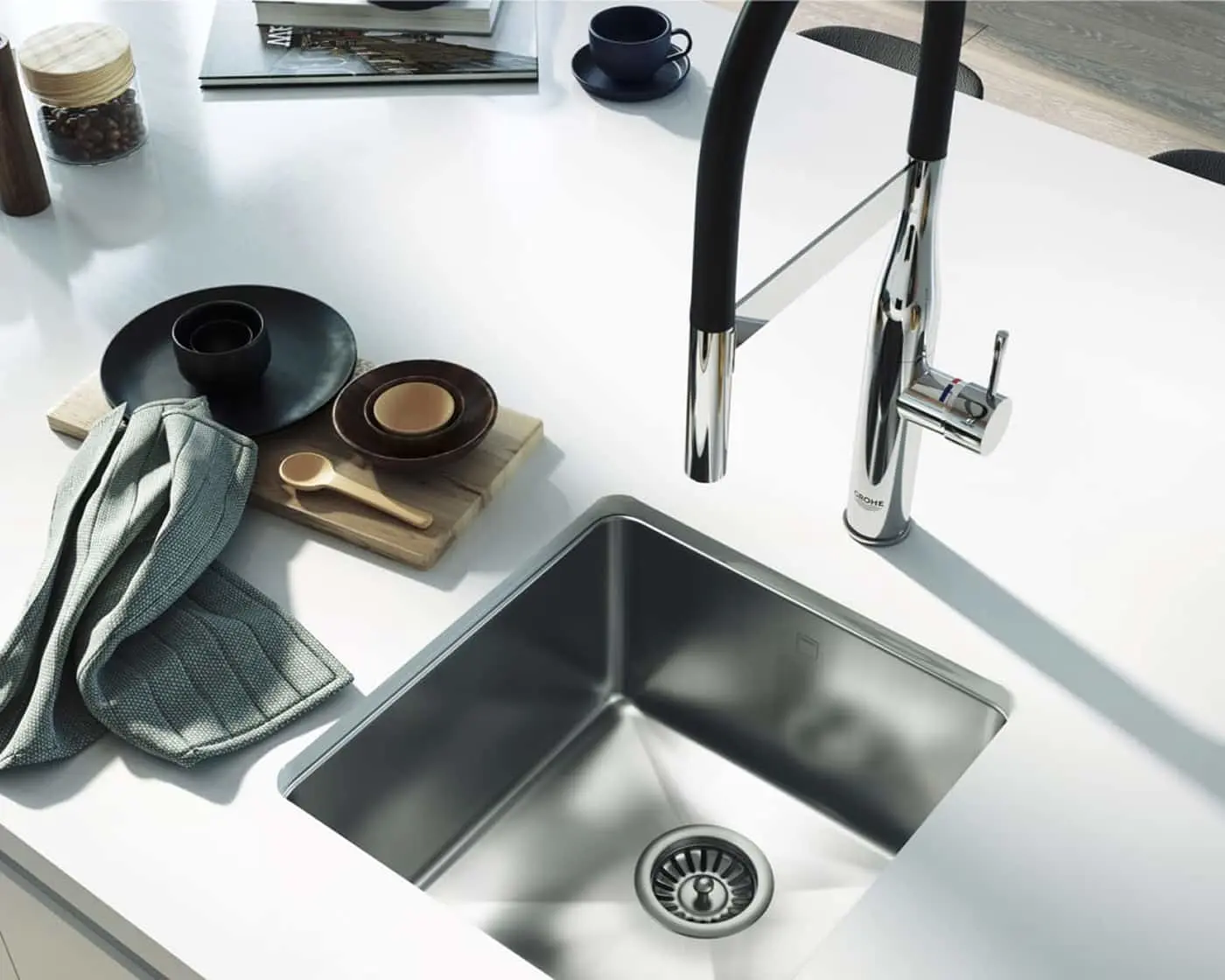 Modern kitchen sink with chrome faucet, white countertop, and dark dishware on wooden board beside blue kitchen towel.