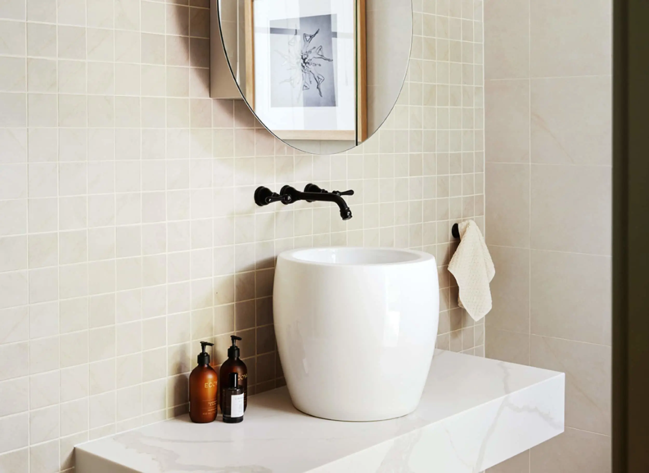 Modern bathroom with white vessel sink, black wall-mounted faucet, round mirror, and beige tiled walls.