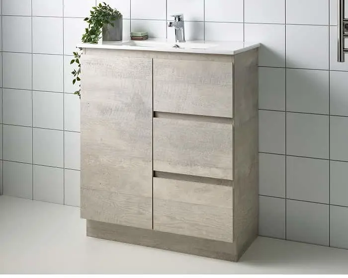 Modern bathroom vanity with light wood finish, white countertop, chrome faucet, and small potted plant against white tiles.